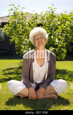 Elder woman sitting on grass with eyes closed in garden outdoors. Meditating in nature in yoga position. Calm and relaxed. Stock Photo