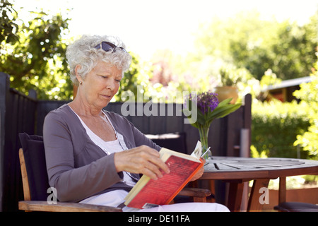 Relaxed senior woman sitting a chair in backyard garden reading a book Stock Photo