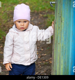 Brown eyed baby girl in pink hat opens old green wooden wicket Stock Photo
