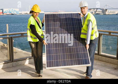 Two people in hard hats and protective jackets carrying a giant solar panel Stock Photo