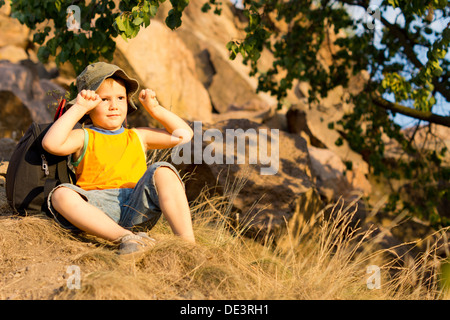Cute little boy sitting on the ground on a mountain slope in his shorts and sunhat flexing his arms muscles with a smile Stock Photo