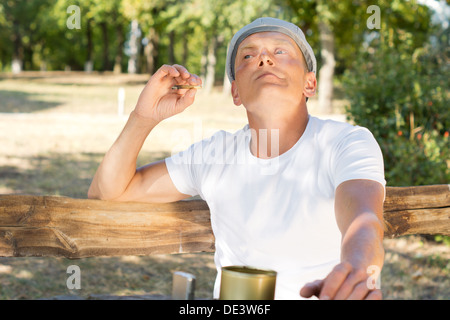 Caucasian man enjoying a rolled cigarette sitting on a bench in the park in a warm sunny day of summer Stock Photo