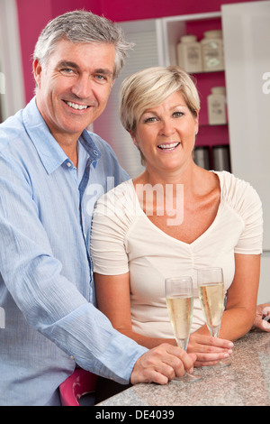 An middle aged happy, smiling man and woman married couple drinking glasses of Champagne in a kitchen at home Stock Photo