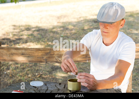 Man rolling himself a cigarette from a small container of fresh tobacco blend while sitting on a wooden park bench Stock Photo