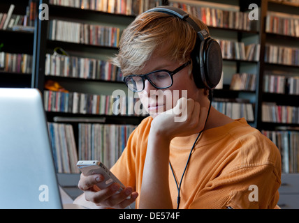 Pensive teenage boy wearing headphones using his smartphone and laptop computer in his home music library Stock Photo