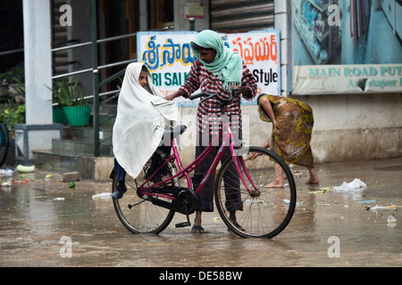 Indian father and son getting on a bicycle in the rain. Puttaparthi, Andhra Pradesh, India Stock Photo