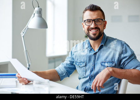Smiling businessman in denim shirt and eyeglasses looking at camera in office Stock Photo