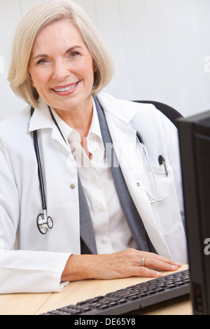 A senior female doctor sitting at a desk in an office with a computer, wearing a white coat and stethoscope Stock Photo