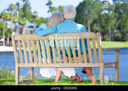 Rear view of a happy romantic senior couple sitting on a park bench embracing looking at a blue lake Stock Photo