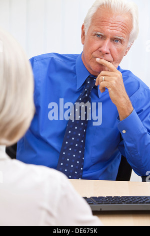 Thoughtful senior businessman sitting at his desk and computer in an office listening to a female colleague opposite him Stock Photo