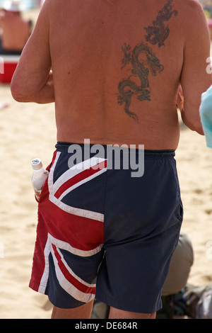 Man with dragon tattoos on back wearing Union Jack shorts with bottle of diet coke in pocket at Bournemouth beach in September Stock Photo