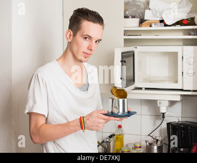 Young serious man standing in a messy kitchen holding plate with metal can Stock Photo