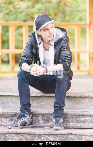 Lifestyle portrait of young man in leather jacket sitting in a park looking away Stock Photo