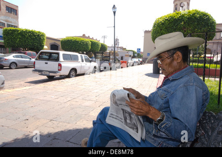 A man wearing a stetson hat reading a newspaper sat on a bench in the shade in Queretaro, Mexico Stock Photo