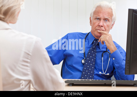 A senior male medical doctor looking thoughtful with a female patient and computer at his desk in an office at a hospital Stock Photo