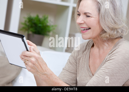 Smiling senior woman at home sitting on sofa or settee using her tablet computer. Stock Photo