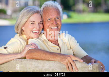 Happy romantic senior couple sitting on a park bench laughing by a blue lake Stock Photo