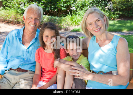 An attractive happy, grandparents, grandson and granddaughter children sitting on a bench outside in the sunshine. Stock Photo