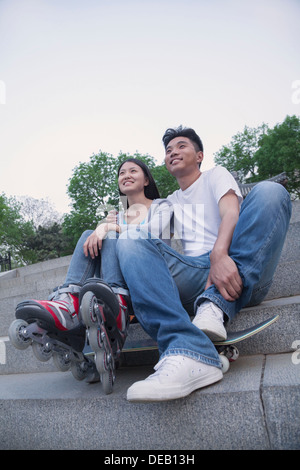 Young happy couple sitting and resting on concrete steps outside with a skateboard and roller blades Stock Photo