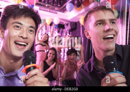 Two friends holding microphones and singing together at karaoke, friends in the background