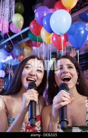 Two friends holding microphones and singing together at karaoke, balloons in the background