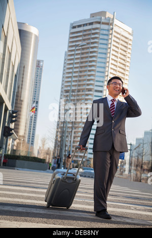 Smiling young Businessman walking down the street with luggage and on the phone Stock Photo