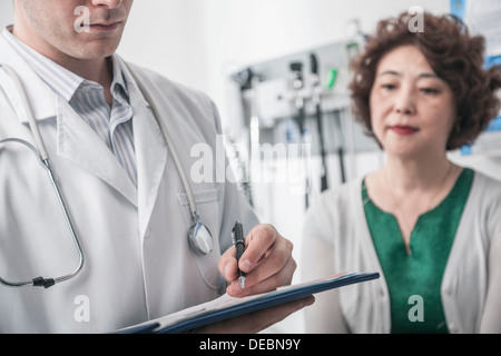 Doctor writing on medical chart with patient in the background Stock Photo