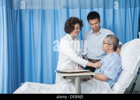 Doctor checking up on patient who is lying down in bed with adult son by his side Stock Photo