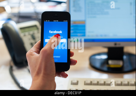 Woman's hand holding a smart phone with facebook on the screen in an office with a desk, phone, computer screen and keyboard Stock Photo