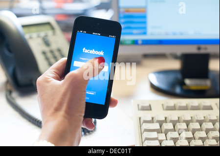 Woman's hand holding a smart phone with facebook on the screen in an office with a desk, phone, computer screen and keyboard Stock Photo