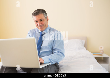 Smiling man using a laptop sitting on bed Stock Photo