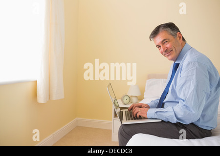 Pleased man using a laptop sitting on a bed Stock Photo