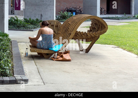 Young woman wearing a sun dress sits studying on a curvilinear wooden bench seat working on her laptop computer. Stock Photo