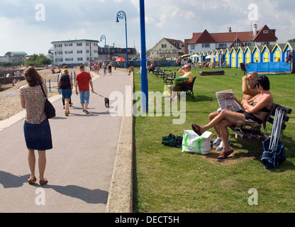 People walk along the seafront while others sit on benches and bask in the sun on the promenade at Felpham in West Sussex, England. Stock Photo