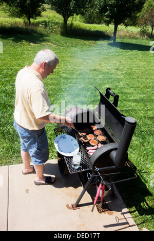 A man in his late 40s grills hamburgers and brats on a smoker/grill in his back yard. Wichita, Kansas, USA. Stock Photo
