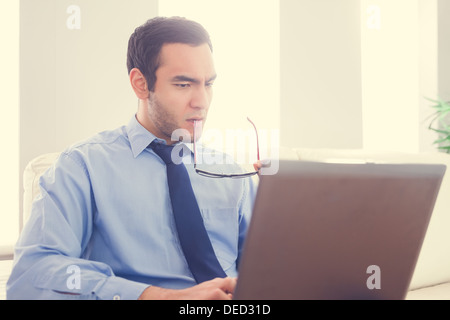Frowning man biting his eyeglasses and using a laptop Stock Photo