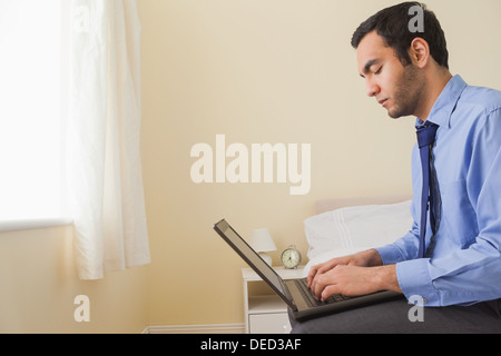 Unsmiling man using a laptop sitting on a bed Stock Photo