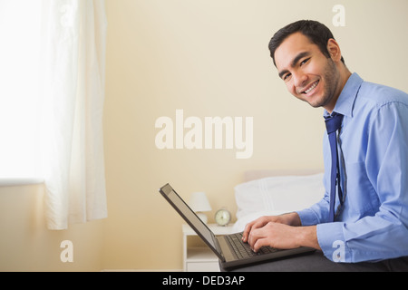 Smiling man using a laptop sitting on bed Stock Photo