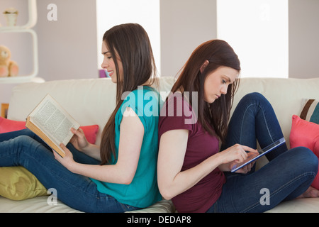 Two happy friends sitting back-to-back on the sofa reading book and using tablet Stock Photo