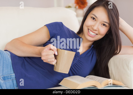 Attractive young asian woman lying on the sofa reading a novel and drinking hot beverage Stock Photo