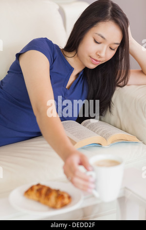 Pretty young asian woman lying on the sofa reading a book reaching for her coffee Stock Photo