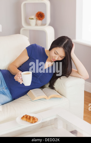 Pretty young asian woman lying on the sofa reading a book holding her coffee Stock Photo