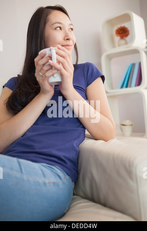 Thoughtful young asian woman sitting on the couch holding mug Stock Photo