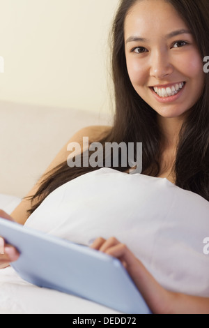Pretty young asian woman sitting in bed using her digital tablet smiling at camera Stock Photo