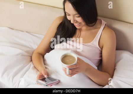Cheerful young asian woman sitting in bed texting on her smartphone and drinking coffee Stock Photo