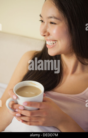 Cheerful young asian woman sitting in bed holding her morning coffee Stock Photo