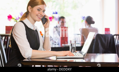 Smiling businesswoman calling on phone typing on laptop and looking at camera Stock Photo