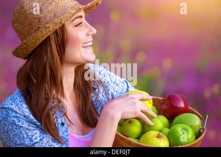 Side view of attractive cheerful female holding in hands basket with apples, fresh ripe fruits, sunset light, pink floral field Stock Photo