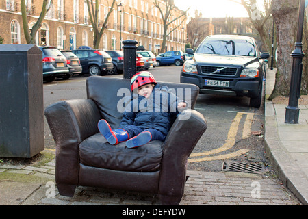 4 year old boy seated in discarded chair on the street Stock Photo