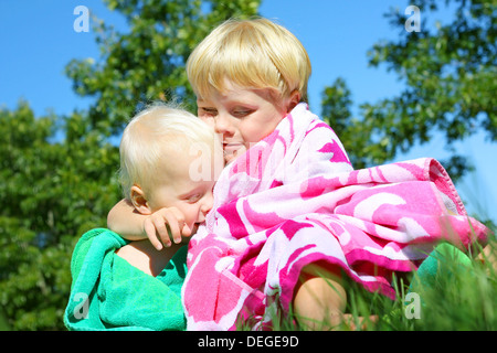 Two happy brothers, a baby and a young child, outside hugging each other in colorful beach towels on a sunny summer day.